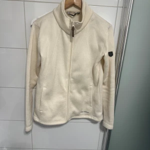 Vit fleecejacka från True North - Säljer en mjuk och varm vit fleecejacka från True North i storlek M. Jackan har hög krage, hel dragkedja framtill och två sidofickor. Perfekt för kyliga dagar och enkel att slänga på över en t-shirt. Stilren design med diskret logga på ärmen.