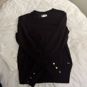 Mörkröd  tröja Holly & Whyte M - Mörkröd långärmad tröja från Holly & Whyte by Lindex i storlek M. Tröjan har rund hals och snygga guldfärgade knappar vid ärmsluten som ger en cool detalj. Materialet är mjukt och känns som bomull, perfekt för chill dagar.