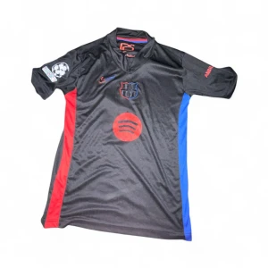 Svart FC Barcelona Pedri 8 matchtröja - Snygg svart FC Barcelona fotbollströja med röda och blå detaljer, Pedri 8 på ryggen och Champions League-märke på ärmen. Spotify-logga på bröstet och klubbmärke i svart och blått. Tillverkad i lätt, ventilerande material som passar perfekt på planen.