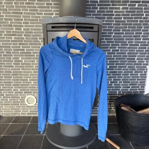 Blå hoodie från Hollister - Snygg blå hoodie från Hollister med vit logga på bröstet och vita snören i huvan. Tröjan har lång ärm och är tillverkad i mjuk bomullsmix, perfekt för en avslappnad stil. Klassisk passform och enkel design som funkar året runt.