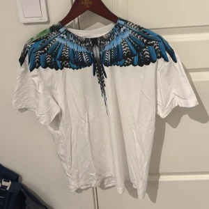 Marcelo Burlon t-shirt med blå fjädrar - Snygg vit t-shirt från Marcelo Burlon County of Milan med ikoniskt blått och svart fjädertryck runt halsen. T-shirten har korta ärmar och är gjord i mjuk bomull. Perfekt statement-plagg för dig som vill sticka ut med cool design. 