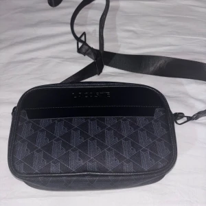 Svart axelväska från Lacoste - Snygg svart axelväska från Lacoste med justerbar rem och diskret logga på både rem och framsida. Väskan har geometriskt mönster och är tillverkad i syntetmaterial med detaljer i skinnimitation. Perfekt storlek för det viktigaste du behöver ha med dig.