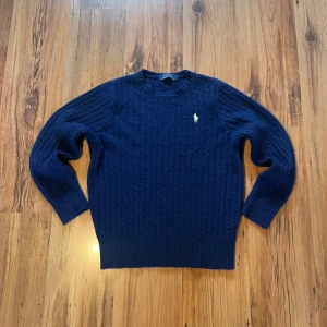 Mörkblå stickad tröja från Polo Ralph Lauren - Kabelstickad mörkblå tröja från Polo Ralph Lauren med klassisk vit broderad logga på bröstet. Tröjan har rund halsringning, långa ärmar och ribbade muddar. Storlekslappen är bortklippt men den sitter som S/170. Längd (60cm), bredd (46cm), ärm (56cm)                Hör av er om ni har frågor 🤩