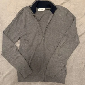 Grå kofta från Pier One - Säljer en stilren grå kofta från Pier One med hel dragkedja och mörkblå krage. Tröjan har långa ärmar och ribbade muddar vid ärmslut och nederkant. Perfekt för lager-på-lager och chill vardagsstil.