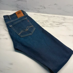 Replay jeans blå tapered fit W29 L32 - Snygga blå jeans från Replay med klassisk femficksdesign och coola kontrastsömmar på bakfickorna. Modellen har tapered fit och är tillverkad i mjukt jeanstyg som sitter skönt hela dagen. Perfekta för dig som gillar stilrena och moderna jeans.