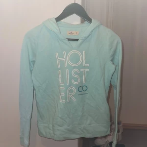 Ljusblå hoodie från Hollister XS - Ljusblå hoodie från Hollister i storlek XS. Tröjan har tryckt logga framtill, huva och långa ärmar. Mjuk och skön bomullsblandning, perfekt för en avslappnad stil. Snygg att matcha med jeans eller leggings.