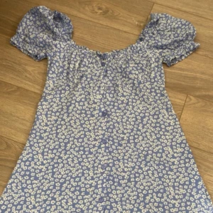 Blommig kortklänning med puffärm - Supersöt kortklänning i ljuslila med vita små blommor och puffiga kortärmar. Klänningen har knappar framtill och en smickrande offshoulder-modell. Perfekt för varma dagar och ger en romantisk vibe.