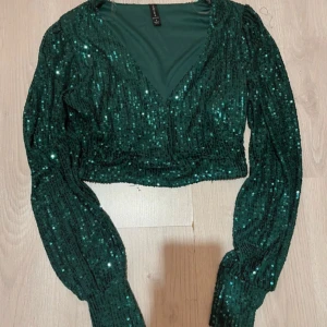 Grön paljett topp med v-ringning - Säljer en glittrig grön croppad topp från lindex med paljetter över hela ytan. Toppen har långärmade puffärmar och djup v-ringning framtill. Perfekt för att sticka ut med extra glam på nästa fest. Supersnygg att matcha med höga jeans eller kjol.