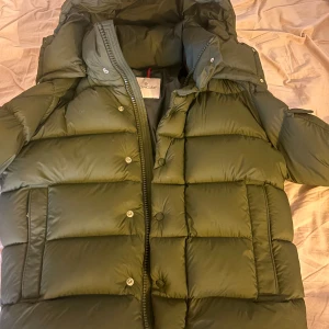 Olivgrön Moncler pufferjacka - Säljer en olivgrön pufferjacka från Moncler med huva och stora, quiltade partier. Jackan har dragkedja och tryckknappar framtill samt en Moncler-logga på ärmen. Perfekt för kalla dagar och riktigt snygg till streetwear. Allt som ingår finns!