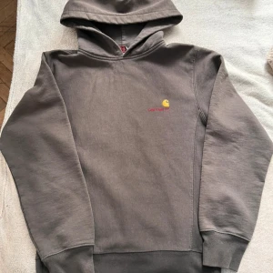 Grå Carhartt hoodie med broderad logga - Säljer en grå hoodie från Carhartt med broderad gul och röd logga på bröstet. Klassisk modell med huva och ribbade muddar. Perfekt för dig som gillar streetwear och vill ha en stilren look.