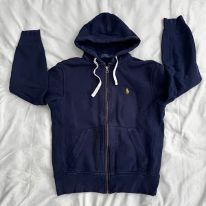 Polo Ralph Lauren zip - Mörkblå zip-hoodie från Ralph Lauren. Tröjan har huva med vita snören, dragkedja & gult märke. Storlek S, passar både man & kvinna.                Nypris: 1200kr. Fraktar inom 24h!