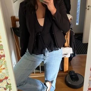 Svart omlottblus med volanger  - En svart omlottblus från Pichnin med v-ringning och volangdetaljer längs axlar och nederkant. Blusen går att ha ”bak och fram” om man vill ha öppen rygg och går även att lätt justera urringning med en liten nål.  