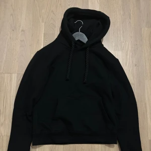 Svart hoodie från Syna World - Svart Syna World hoodie i riktigt snygg streetwear-stil. Hoodien har den ikoniska broderade Syna World-loggan på bröstet och är gjord i tjock, mjuk premiumkvalitet med en skön oversized passform. Perfekt både till vardags och när du vill ha en avslappnad men stilren look.