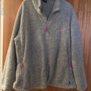 Grå teddyjacka On The Peak XL - Mjuk och fluffig grå teddyjacka från On The Peak i storlek XL. Jackan har hel dragkedja framtill och två sidofickor med rosa detaljer på dragkedjorna. Perfekt för kyliga dagar och har en avslappnad passform.