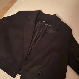 Svart kavaj från H&M, storlek 38 - Snygg svart kavaj från H&M i klassisk modell med två knappar framtill och smal krage. Kavajen har en stilren look och är fodrad med svart tyg på insidan. Perfekt för dig som gillar en enkel och clean stil. Aldrig använt och det vita e liksom nåhot från luften det tar man bort lätt. Köpte för 500 kr 