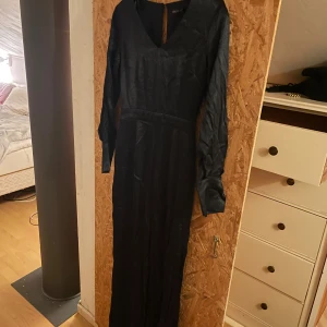 Svart jumpsuit med v-ringning - Snygg svart jumpsuit från  Stockholm med v-ringad hals och långa ärmar. Klänningen har en elegant, enkel design och är tillverkad i ett glansigt material som ger en lyxig känsla. Perfekt för dig som vill sticka ut med stil.