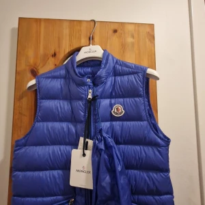 Blå dunväst från Moncler - Snygg blå dunväst från Moncler med klassisk quiltad design och dragkedja framtill. Västen har hög krage och ikonisk Moncler-logga på bröstet. Perfekt för lager-på-lager och ger en sportig vibe till din outfit.