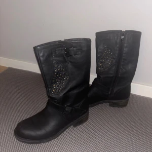Svarta boots med nitar och spännen -  Priset går att diskuteras !Säljer ett par svarta boots i skinnimitation med coola nitar på sidorna och snygga spänndetaljer upptill och vid ankeln. Dragkedja på insidan för enkel påtagning. Perfekta för dig som vill ha en edgy stil till hösten eller vintern.