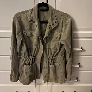 Grön  parkajacka från Gina Tricot - Säljer en grön parkajacka från Gina Tricot i storlek 36. Jackan har snörning i midjan, flera fickor framtill och knappar längs öppningen. Materialet är bomull och jackan har en cool, lite loose passform med långärmade ärmar.