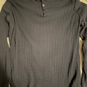 Mörkblå stickad tröja från Mywear XL - Mörkblå stickad tröja från Mywear i storlek XL. Tröjan har ett klassiskt kabelstickat mönster, lång ärm och knäppning vid halsen. Perfekt för kyligare dagar och enkel att matcha med jeans eller chinos.