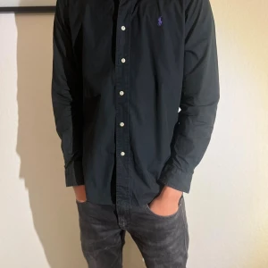 Svart skjorta från Ralph Lauren - Svart långärmad skjorta från Ralph Lauren med klassisk krage och vita knappar framtill. Diskret lila logga broderad på bröstet. Skjortan är i bomull och har en normal passform som sitter snyggt. Perfekt för dig som gillar stilrena och tidlösa plagg.