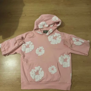 Rosa hoodie från Denim Tears med blommor - Säljer en rosa hoodie från Denim Tears med vita blommiga tryck över hela tröjan. Hoodien har huva, magficka och ribbade muddar vid ärmslut. Tillverkad i USA och gjord i mjuk bomull, perfekt för dig som gillar unik streetwear.