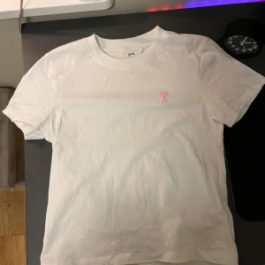 Vit t-shirt från Ami  - Vit t-shirt från Ami med klassisk passform och rund hals. På bröstet finns en broderad rosa hjärtdetalj med bokstaven A. Tillverkad i mjuk bomull som känns skön mot huden. Perfekt för en clean och stilren look. Helt oanvänd och ny! Pris kan diskuteras!
