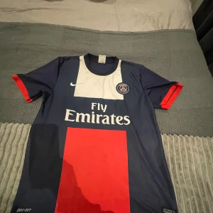 PSG Ibrahimović 10 Nike fotbollströja - Paris Saint-Germain matchtröja med Ibrahimović 10 på ryggen. Tröjan är marinblå med röd och vit detalj, klassisk Nike Dri-FIT med korta ärmar och klubbmärke på bröstet. Snygg design med Fly Emirates-tryck framtill och röda muddar.