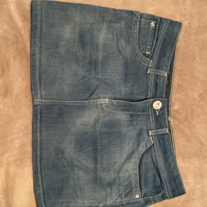 Blå jeanskjol  - Snygg blå jeanskjol med klassisk femficksdesign, vita kontrastsömmar och dragkedja framtill. Kjolen är kort och har bälteshällor samt dekorativa bakfickor. Perfekt för dig som gillar en enkel och tidlös denimlook.