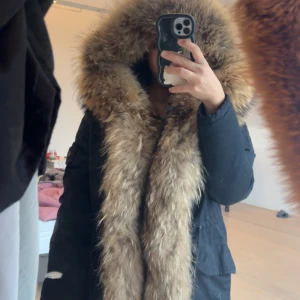 Svart parkajacka med stor päls från The Smirk - Parkajacka från The Smirk med massiv fluffig päls i bruna toner runt huvan och längs öppningen. Jackan är svart, har snörning i midjan och långa ärmar. Perfekt för dig som vill ha en statement vinterjacka med extra mycket pälsdetaljer. Äkta päls kan tänka mig gå ner i pris vid snabb affär 