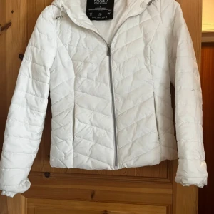 Vit quiltad pufferjacka Stradivarius - Snygg vit pufferjacka från Stradivarius med huva och dragkedja framtill. Jackan har ett quiltat mönster i chevronstil och är tillverkad i lätt polyester. Perfekt för dig som vill ha en clean och fräsch look under kyliga dagar. Storleken är M men funkar mest för S (liten i storlek)