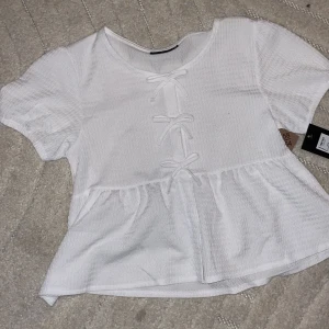 Vit blus med rosetter från XOXO Friends - Supersöt vit blus från XOXO Friends i storlek 158/164. Blusen har korta puffärmar, rund halsringning och tre dekorativa rosetter framtill. Materialet är lätt strukturerat och ger en mjuk känsla. Perfekt för dig som gillar gulliga detaljer och en feminin look.