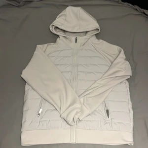 Gråvit pufferjacka/cardigan med huva och dragkedja - Snygg gråvit pufferjacka med huva och hel dragkedja framtill. Jackan har quiltad front och rygg, två sidofickor med dragkedja och mjuka långärmade ärmar. Perfekt för dig som gillar stilren och clean look. Ej använd då den inte passa mig, pris kan diskuteras