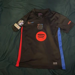FC Barcelona Lamine Yamal Nike tröja S - Säljer en svart FC Barcelona fotbollströja från Nike med Lamine Yamal och nummer 19 på ryggen. Tröjan har röda och blå detaljer, Champions League-märke på ärmen, Spotify-logga på bröstet och UNHCR-tryck bak. Tillverkad i Dri-FIT-material för bästa komfort.