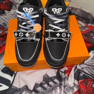 Louis Vuitton LV Trainer sneakers svart - Säljer ett par Louis Vuitton LV Trainer sneakers i svart med vita detaljer och grå sula. Skorna har klassiska LV-loggor, monogramdetaljer och snörning samt kardborreband med ikoniska symboler. Materialet är en mix av skinn och textil för en lyxig känsla.