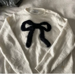 Vit stickad tröja med svart rosett - Superfin vit stickad tröja från Holly & Whyte med en stor svart fluffig rosett framtill. Tröjan har rund halsringning, långa ärmar och ribbade muddar. Perfekt för dig som gillar söta detaljer och vill ha en mysig look.