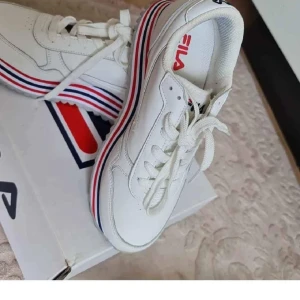 Vita Fila sneakers med rödblå rand - Snygga vita Fila sneakers med platt sula och coola röda och blå ränder längs sulan. Skorna har snörning och rund tå, samt Fila-logga på både häl och plös. Perfekta för dig som gillar sportig och fräsch stil.