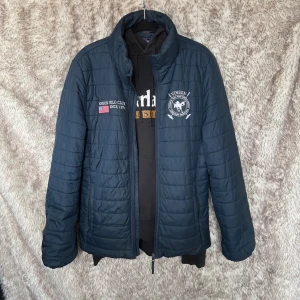 Mörkblå quiltad jacka Vinson Polo - Quiltad mörkblå jacka från Vinson Polo Club med broderade loggor och amerikansk flagga på bröstet. Båda dragkedjorna vid sidofickorna har hoppat ur spåren. Annars väldigt fin.