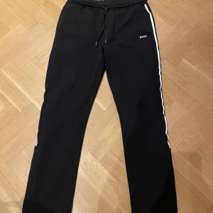 Svarta mjukisbyxor från BOSS XL - Svarta raka mjukisbyxor från BOSS i storlek XL med vita ränder längs sidorna och diskret BOSS-logga på benet. Byxorna har elastisk midja med snörning och är tillverkade i mjukt material som känns skönt mot huden. Perfekta för en chill och sportig look. Använd fåtal gånger och därmed i bra skick. 