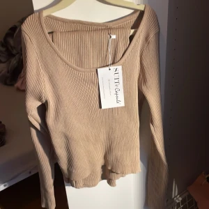 Beige ribbad croppad tröja Suite Capsule med en matchande lång kjol med slits  - Snygg beige croppad tröja från Suite Capsule med ribbad struktur och lång ärm. Tröjan har en rundad halsringning och cool slitsdetalj nedtill. Tillverkad i mjuk viskosblandning som sitter skönt mot kroppen. Perfekt för dig som gillar stilrena och trendiga plagg.