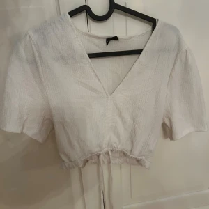 Vit croppad blus med snörning - Supersöt vit blus med v-ringning och korta puffärmar. Toppen är croppad med snörning i midjan och har en lätt textur i tyget 