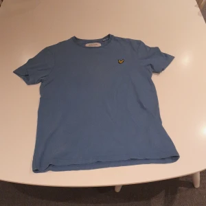 Blå t-shirt från Lyle & Scott XS - Snygg blå t-shirt från Lyle & Scott i ekologisk bomull. Klassisk passform med rund hals och korta ärmar. På bröstet sitter den ikoniska gula örnloggan broderad. Perfekt för en clean och stilren look.