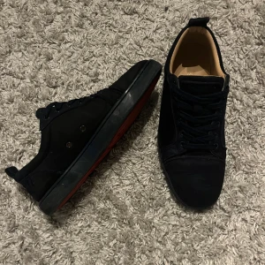 Mörkblåa Christian Louboutins - Snygga svarta sneakers med rund tå och platt sula. Skorna har snörning och en cool röd undersida med mönstrad design. Utsidan är i mocka och insidan är ljusbeige. Perfekta för dig som gillar stilrena och unika detaljer.