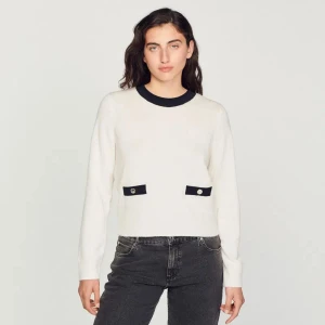 Vit tröja från Sandro med svarta detaljer - Vit tröja från Sandro med svarta kontrastdetaljer vid halsringning, fickor och längs ryggen. Guldfärgade knappar både fram och bak. Material: 94% ull, 6 % Kashmir gör att den är varm och skön. Enbart använd två gånger- som ny! Nypris: 2900 kr