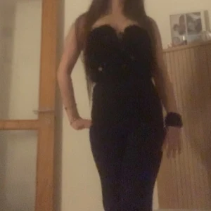 Svart byxdress med paljetter - Snygg svart byxdress med figurnära passform och utsvängda ben från Newyorker ((ANVÄND BARA EN GÅNG)). Överdelen är axelbandslös och dekorerad med paljetter och fluffiga detaljer för en extra festlig look. Perfekt för dig som vill sticka ut.  Säljer den eftersom den var för lång och stor på mig. 