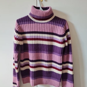 Varm ulltröja från Åhléns strl L - Säljer en ribbstickad polotröja retro från Åhléns i storlek L, gjord av 70% lammull och 30% akryl. Tröjan har breda ränder i lila, rosa, vinröd och vit, samt långärmad design och hög krage.