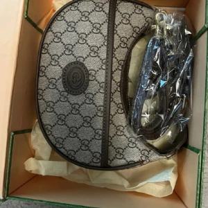 Gucci GG Supreme axelväska brun - Gucci axelväska i klassiskt GG Supreme-mönster med bruna skinn-detaljer och guldfärgad dragkedja. Väskan har halvmåneform och justerbar axelrem. Insidan är fodrad med beige tyg och har präglad Gucci-logga. Perfekt för dig som gillar lyxiga accessoarer.