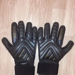 Adidas Copa målvaktshandskar svart/guld - Snygga Adidas Copa Pro målvaktshandskar i svart med guldiga detaljer och Adidas-logga på fingrarna. Handskarna har en sportig design med vaddering och breda muddar med kardborre. Perfekta för fotbollsmålvakter som vill ha stil och grepp på planen.