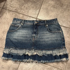Jeanskjol med fransad volangkant - Cool jeanskjol i mörkblå denim med slitna detaljer och fransad volang längst ner. Kjolen har klassiska fickor fram och bak, bälteshällor och knappstängning. Perfekt för dig som gillar en edgy och trendig stil.