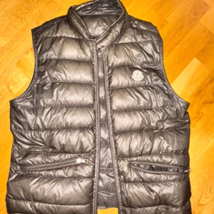 Svart dunväst från Moncler - FINNS I STOCKHOLM! Snygg svart dunväst från Moncler med glansig finish och klassisk logga på bröstet. Västen har ståkrage, två dragkedjefickor framtill och quiltad design. Perfekt för lager-på-lager och ger en clean streetstyle-vibe.